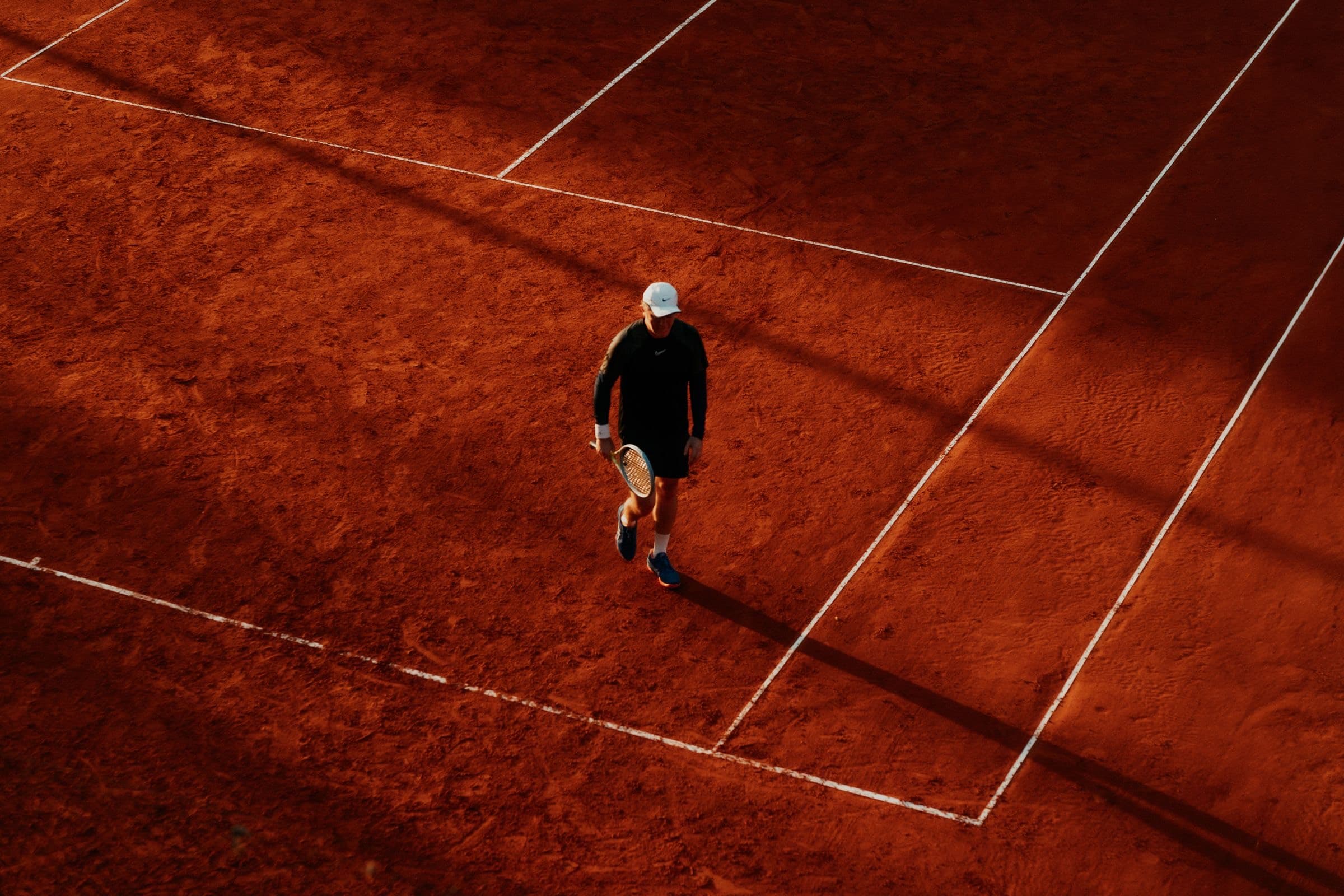 A player walking across a clay court in late sun, seen from above